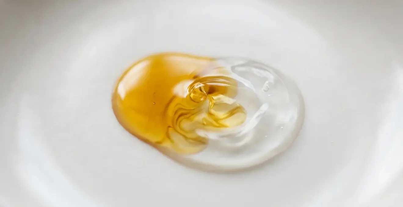 Gros plan macro sur deux textures de nettoyants, huile dorée et gel translucide, se mélant délicatement